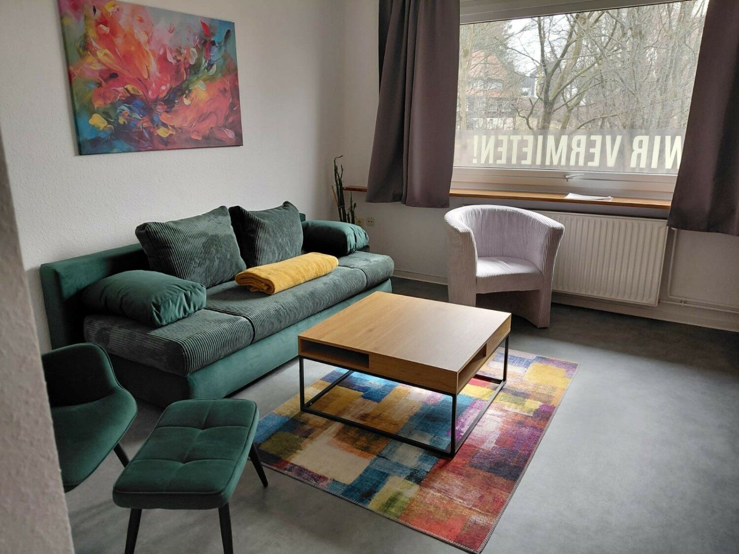 Wohnzimmer mit grünem Sofa, Sessel und buntem Teppich